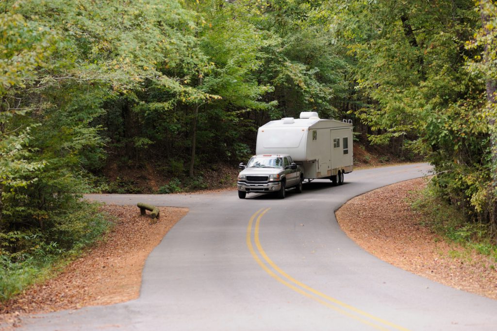 Recreational vehicle fifth wheel travel trailer being towed down curving road in woods.